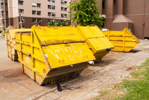 A large yellow industrial skip bin with rusted metal surfaces and reinforced sides, positioned on a paved area near a multi-storey residential building. The skip is partially tilted, with its open top showing signs of dirt and wear, and the side panels have black markings and weathering. The bin's edges feature metal reinforcements, and the front is slightly raised off the ground, revealing a dark interior. Surrounding the skip are patches of grass and a narrow sidewalk, with an urban apartment complex visible in the background, including balconies and windows. The scene is illuminated by daylight, creating shadows around the bin, which is likely used by Commercial Waste Removal Anerley for rubbish collection and disposal in the local area post code area. The overall environment suggests a temporary storage spot for waste materials to be removed in accordance with local waste management services.