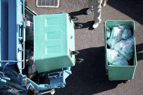 An aerial view of a teal waste collection trolley positioned next to a green plastic bin filled with mixed waste, including plastic bags and other refuse, on a paved outdoor surface. A person wearing light-colored clothing and white shoes stands nearby, partially visible in the upper part of the image. In the background, part of a waste collection vehicle with a blue exterior is visible, indicating a commercial rubbish removal activity. The environment appears to be a typical outdoor setting associated with waste management in Anerley, with the scene captured in daylight providing natural illumination.