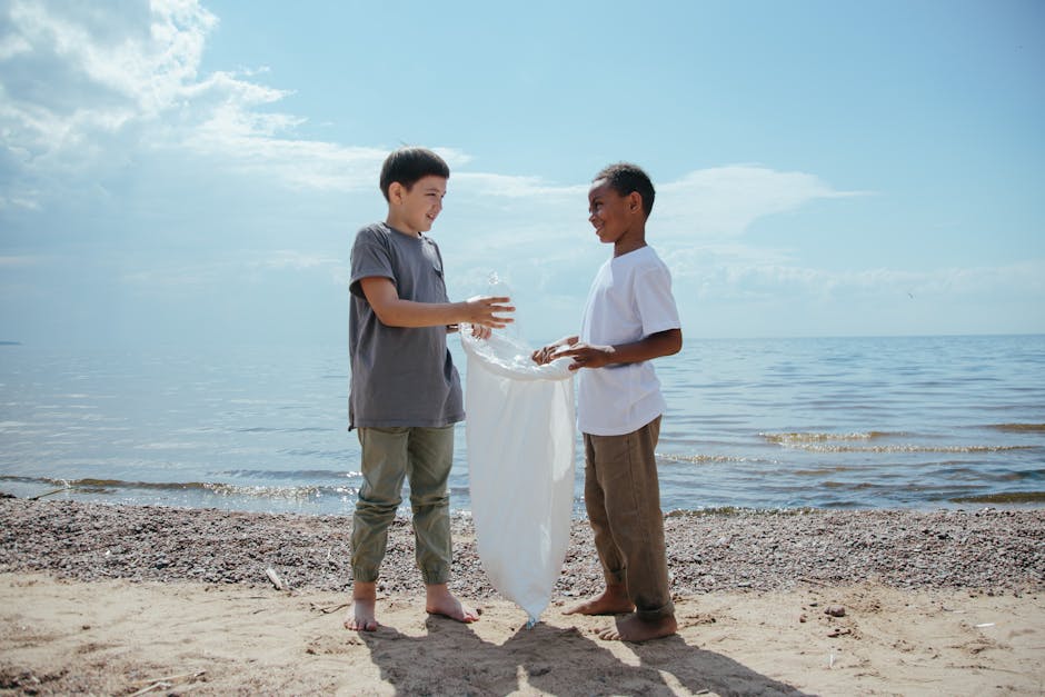 Two boys stand barefoot on a sandy beach near the shoreline, engaging in a recycling activity by exchanging a clear plastic bottle and placing it into a white fabric bag held between them. The boy on
