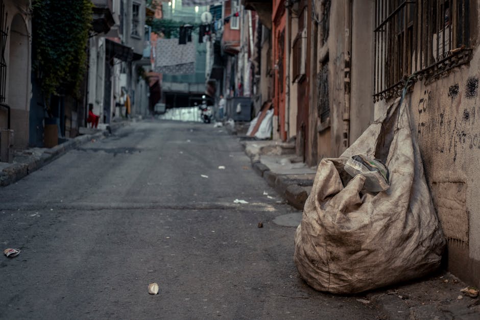 A large, dirty, beige cloth sack filled with trash and debris is positioned on the sidewalk next to an old, weathered brick wall with black wrought iron window bars in a narrow urban street. The sack 