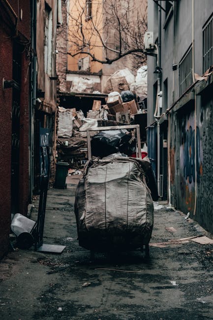 A large, dirty, beige cloth sack filled with trash and debris is positioned on the sidewalk next to an old, weathered brick wall with black wrought iron window bars in a narrow urban street. The sack 