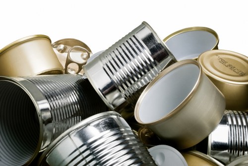 A close-up of a collection of discarded metal containers and cans, including both aluminium and tin, stacked and scattered on a surface. The cans vary in size, some with open tops revealing smooth, reflective interior surfaces, while others remain closed with rolled edges or stamped lids. The metallic textures display a shiny, reflective finish, with some showing slight ridges from manufacturing. The background is neutral and out of focus, emphasizing the clutter of scrap metal items typically encountered during rubbish removal in commercial or industrial settings. The arrangement offers a detailed view suitable for illustrating waste management or rubbish removal services in Anerley, with the presence of these recyclable materials highlighting the importance of proper disposal and collection by companies like Commercial Waste Removal Anerley.