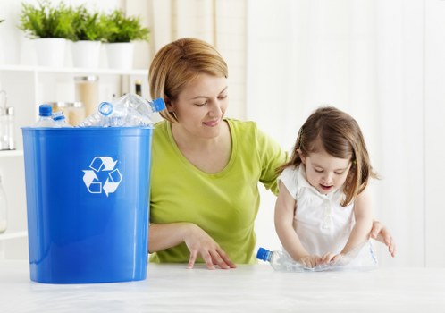 A woman with short, light brown hair wearing a bright yellow-green top is seated at a table indoors with a young girl who has shoulder-length brown hair dressed in a white top. They are engaged in a recycling activity, with the woman gently guiding the girl as she places a clear plastic bottle into a blue recycling bin that features a white recycling symbol on its front. The bin is positioned on the left side of the image, and it is filled with crushed or empty plastic bottles made of transparent PET material, some with blue caps. The background shows a well-lit, modern interior with white shelving units holding neatly arranged potted plants, contributing to a clean and orderly environment. The scene captures a moment of environmental awareness focusing on waste separation, with the emphasis on plastic waste disposal, aligning with rubbish removal services in Anerley, near London, where local waste management practices are integral to community sustainability efforts.