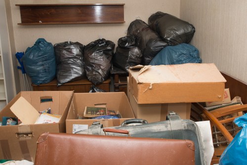 The image shows a cluttered indoor space, possibly a storage room or corner, filled with various bags and boxes of waste items. In the background, there are seven large black waste bags, some tied at the top, arranged on a wooden shelf against a beige wall. To the right of these, a couple of blue rubbish bags rest directly on the shelf. In the foreground, there are several open cardboard boxes, one containing papers, and a metallic object resembling a compressed gas cylinder or portable heater, partially visible. A large, flat, light brown cardboard box with a torn corner sits atop the boxes, and an orange-brown piece of furniture or cushion is partially visible at the bottom front of the image. The overall setting suggests a typical household clutter area awaiting rubbish removal, and the space is lit by natural or ambient light, reflecting a functional but disorganized environment, typical of waste clearance activities by companies such as Commercial Waste Removal Anerley, servicing areas around Anerley and the postcode district.